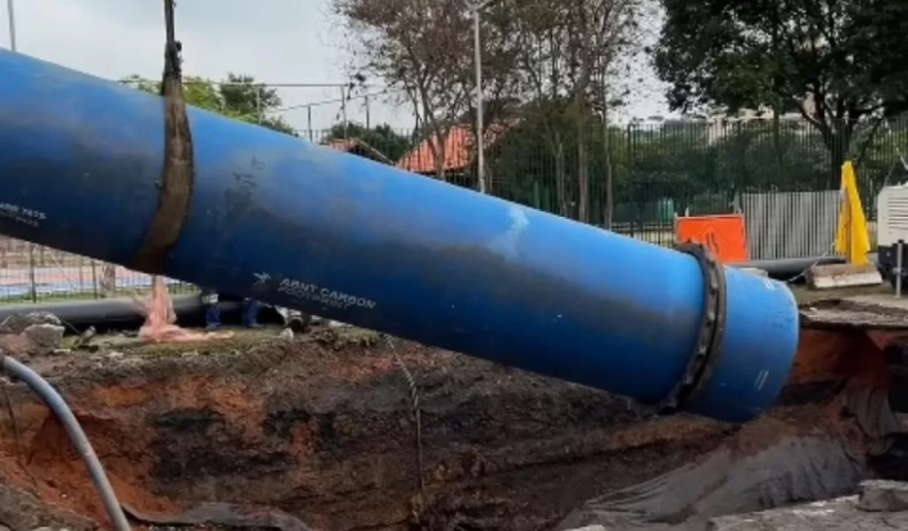 Um grande tubo azul de infraestrutura é içado por cintas sobre uma vala profunda em um canteiro de obras urbano, cercado por árvores e sinalizações de segurança.
