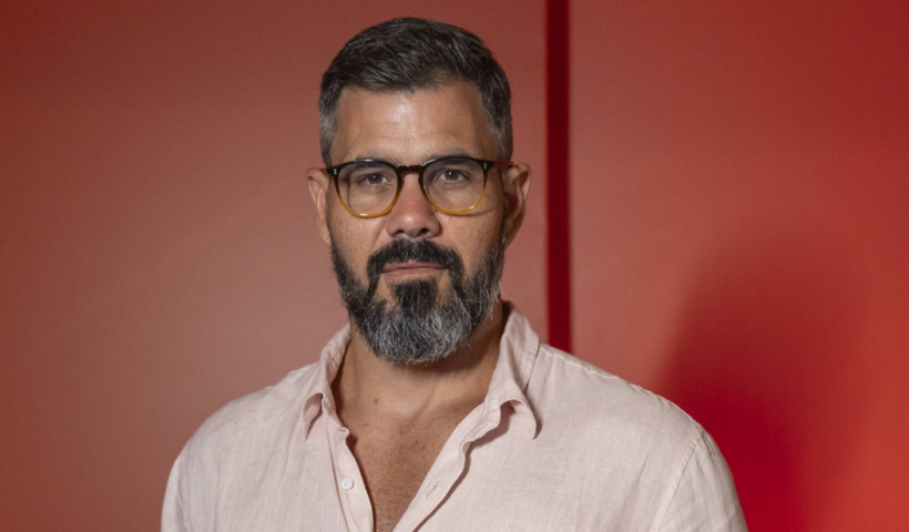 Ator Juliano Cazarré com camisa rosada, olhando sério para câmera