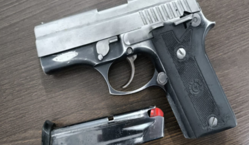 Uma pistola semiautomática com ferrolho prateado e empunhadura preta está posicionada sobre uma superfície de madeira ao lado de seu carregador desencaixado