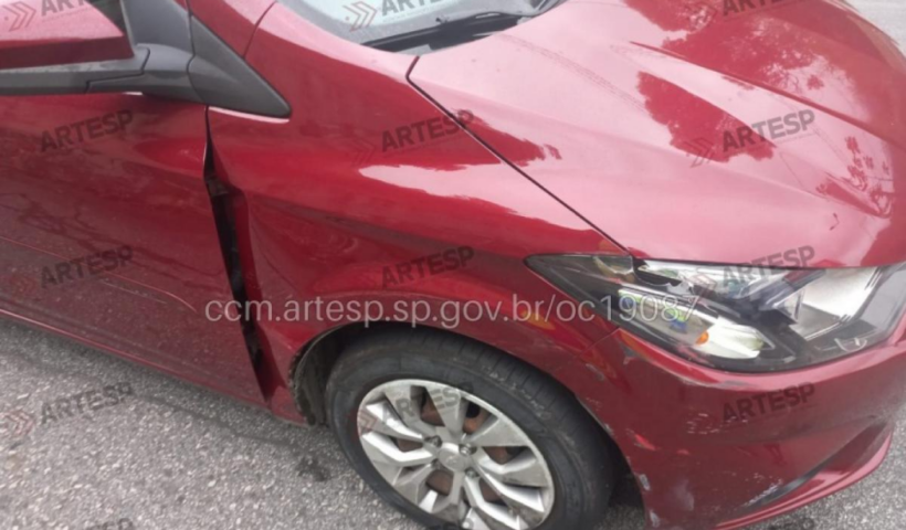 A imagem mostra a lateral dianteira de um carro vermelho com o para-lama amassado, o farol quebrado e o pneu dianteiro direito completamente murcho após um impacto.