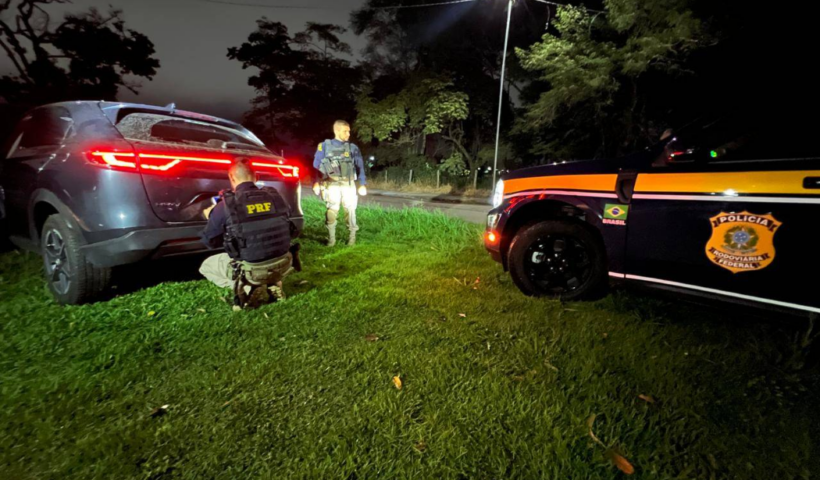 À noite, na beira de uma estrada, dois agentes da Polícia Rodoviária Federal (PRF), com distintivos visíveis em seus coletes e viatura, fiscalizam um SUV cinza parado na grama, com as lanternas traseiras acesas.