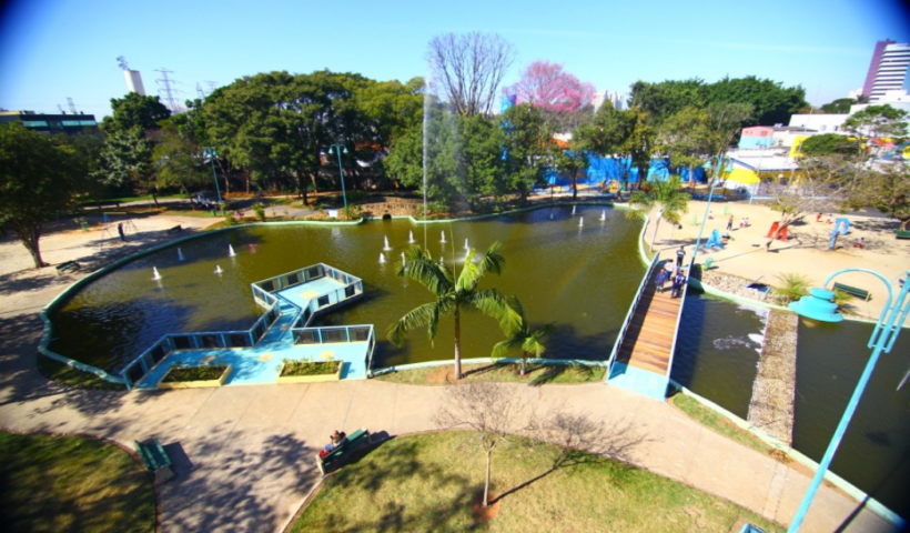 Vista aérea do Parque Santos Dumont revela um lago central com chafarizes, pontes e plataformas azuis, cercado por gramados, árvores frondosas e caminhos pavimentados sob um céu ensolarado.