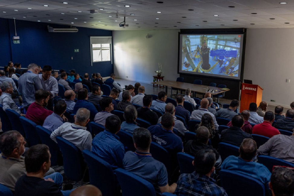 auditório com funsionários da avibras e sindicalistas; no palco dp auditório tem uma mesa de reunião e um telão para vídeos ao fundo