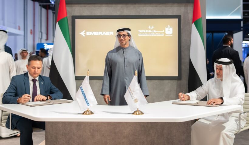 representante da Embraer à esquerda, representante da Tawazun Council for Defence Enablement, sentados à mesa assinando arcodo comercial; outro representante dos Emirados árabes ao centro, de pé, sorrindo para a foto