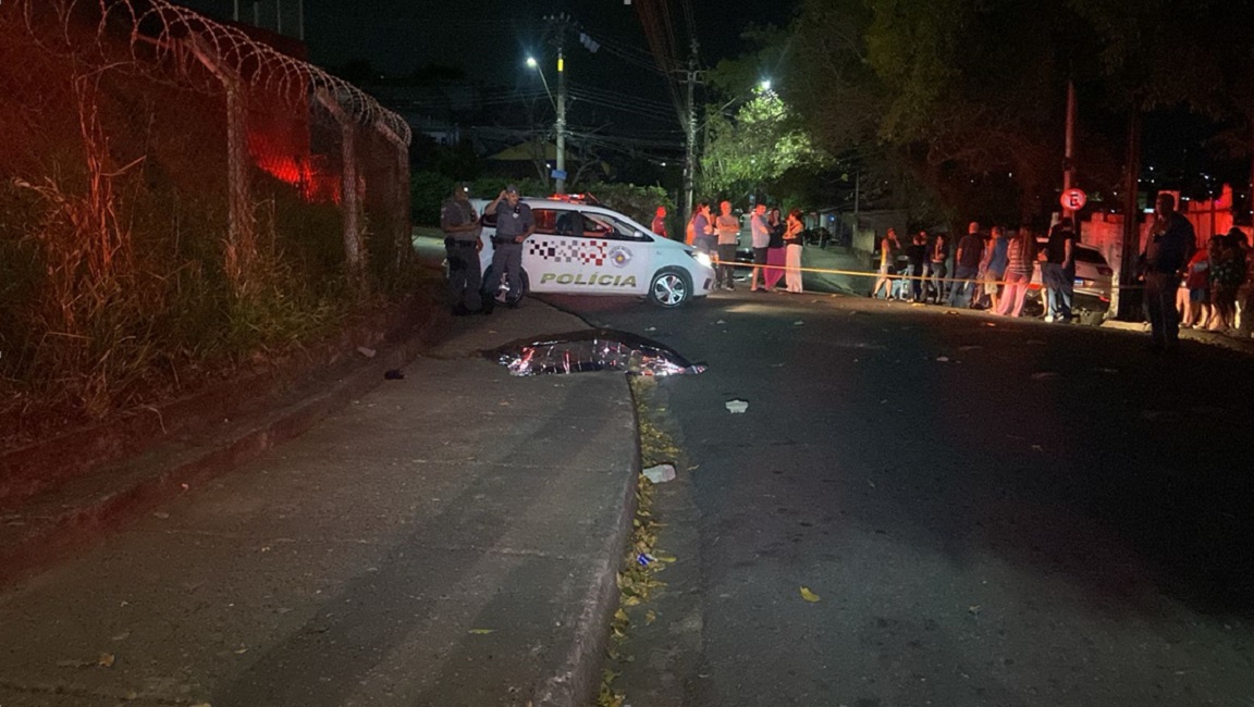 corpo de homem vítima de execução, coberto por papel alumínio, ao lado de viatura da política; ambiente noturno
