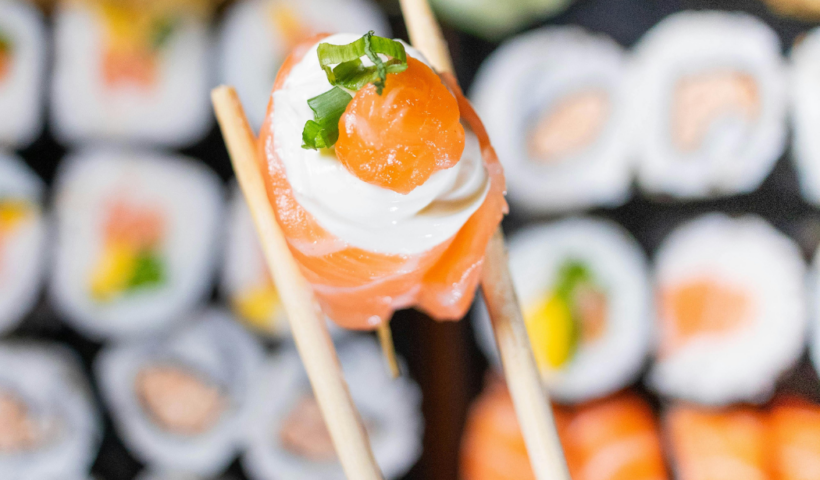 Um par de hashis segura em destaque um sushi jojo de salmão com cream cheese e cebolinha, tendo ao fundo diversos outros sushis variados desfocados.