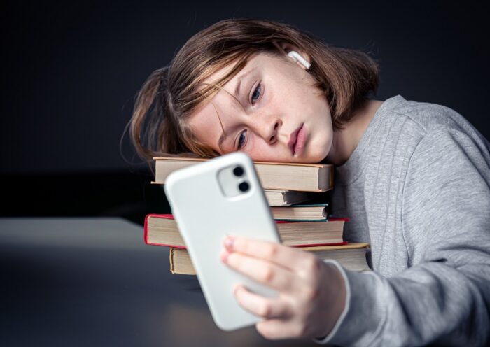 Adolescente sentada com livros, mas usando um smartphone; tédio com a leitura e dependência de tecnologia; Câmara aprova projeto sobre nomofobia em São José dos Campos; entenda
