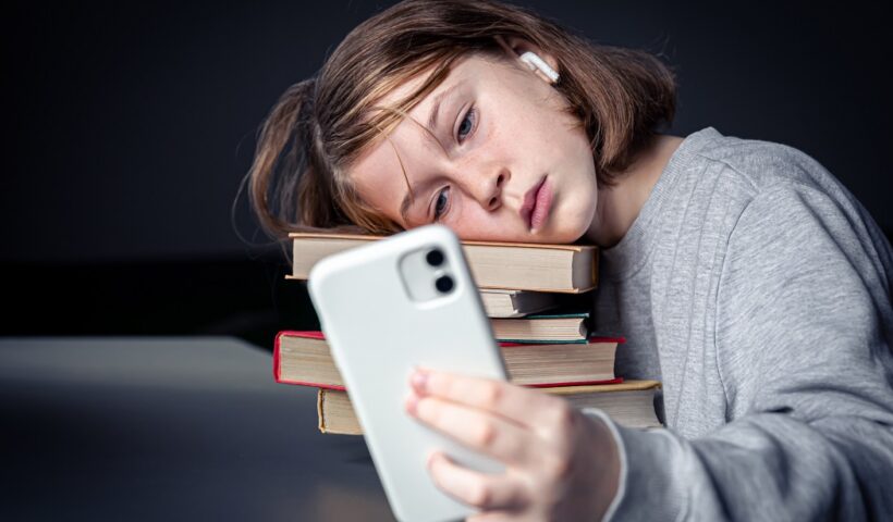 Adolescente sentada com livros, mas usando um smartphone; tédio com a leitura e dependência de tecnologia; Câmara aprova projeto sobre nomofobia em São José dos Campos; entenda