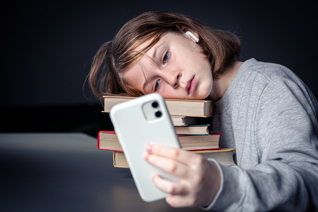 Adolescente sentada com livros, mas usando um smartphone; tédio com a leitura e dependência de tecnologia; Câmara aprova projeto sobre nomofobia em São José dos Campos; entenda